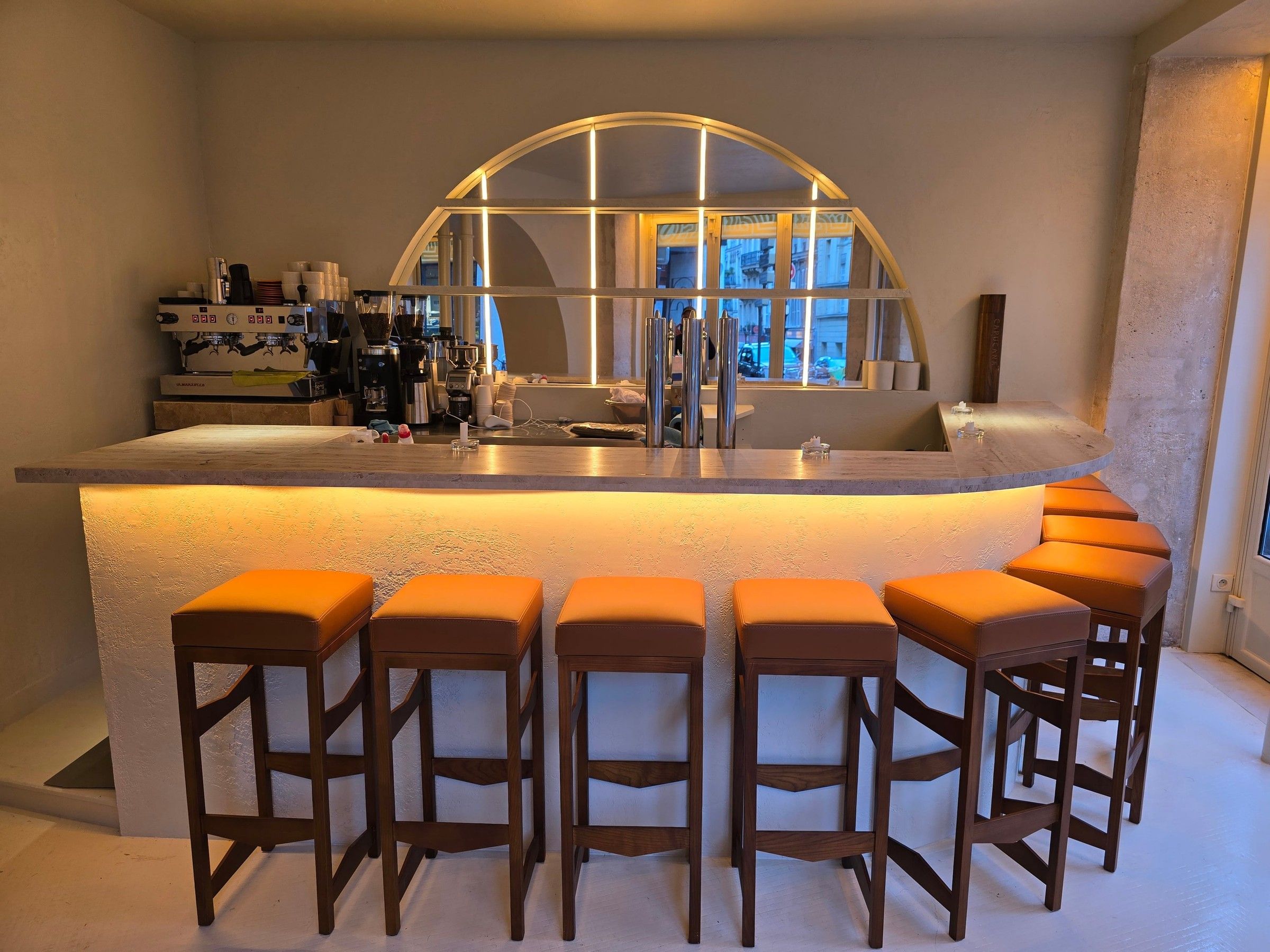 Bar vu de face, éclairé sous le plateau en pierre, avec une rangée de tabourets en bois à assise camel