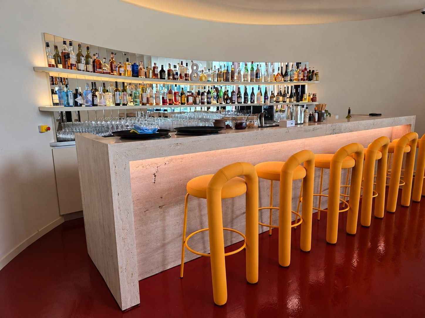 Bar en pierre claire rétroéclairé et tabourets jaunes, devant l'étagère à bouteilles